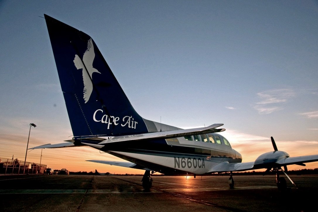 Cessna blanc de Cape Air avec empennage arrière bleu décoré d'une colombe blanche, pris de l'arrière, sur la piste avec le coucher de soleil en arrière-plan.