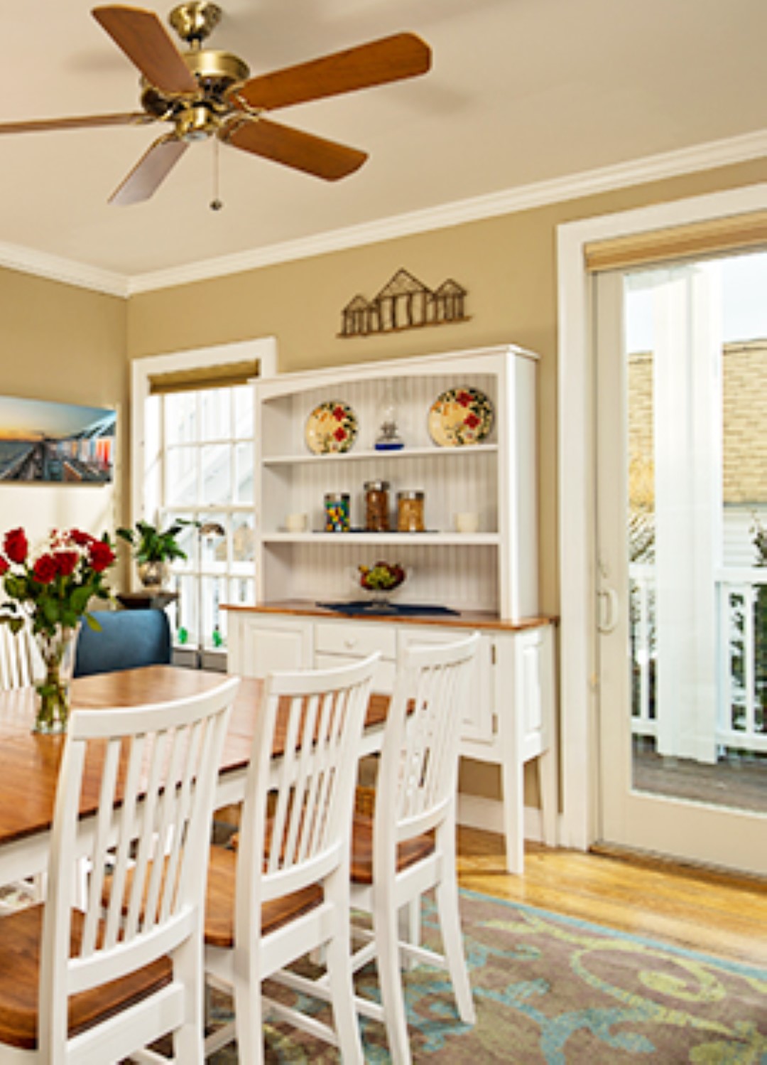 Salle à manger avec table et chaises peintes en blanc avec plateau en acajou, buffet blanc avec trois étagères en arrière-plan entre une fenêtre à gauche et une grande porte coulissante donnant sur la terrasse à droite.